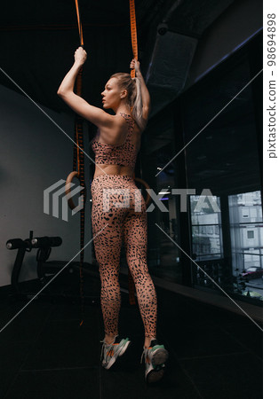 Gimnastic rings. Muscular back of a girl Crossfit athlete in black sportswear is hanging on gymnastic rings. Caucasian woman with long blond hair training olympic ring. Gimnastic rings. Muscular back of a girl Crossfit athlete in black sportswear is hanging on gymnastic rings. Caucasian woman with long blond hair training olympic ring. 98694898