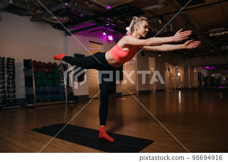 Beautiful young blond girl yoga instructor doing on a mat in a floor standing in the gym. Young sport woman standing on one leg and exercising. 98694916