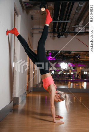 Young woman in sportswear exercising yoga asana in handstand leaning legs on wall. Woman practicing yoga on floor in headstand pose upside down balancing body leaning wall at fitness studio. 98694922