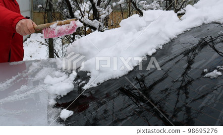 a man in a red jacket cleans the windshield of a car with a reflection of branches on the glass from a layer of snow using a home brush, removing snow from the body of a private car in the yard a man in a red jacket cleans the windshield of a car with a reflection of branches on the glass from a layer of snow using a home brush, removing snow from the body of a private car in the yard 98695276