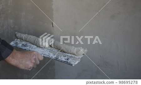 a plasterer's hand with a wide spatula and a thick elastic mortar putty and level the wall and copy space on the left on a gray cement surface, an example of plastering a room wall a plasterer's hand with a wide spatula and a thick elastic mortar putty and level the wall and copy space on the left on a gray cement surface, an example of plastering a room wall 98695278