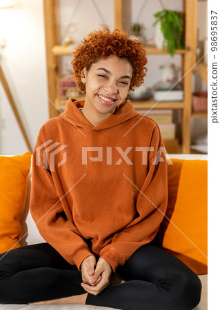 Beautiful african american girl with afro hairstyle smiling sitting on sofa at home indoor. Young african woman with curly hair laughing. Freedom happiness carefree happy people concept Beautiful african american girl with afro hairstyle smiling sitting on sofa at home indoor. Young african woman with curly hair laughing. Freedom happiness carefree happy people concept 98695837