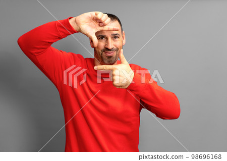 Handsome bearded happy male blinks his eyes and looks through a frame formed by his hands. An attractive man makes a frame with his fingers, isolated on a gray background with copy space for text Handsome bearded happy male blinks his eyes and looks through a frame formed by his hands. An attractive man makes a frame with his fingers, isolated on a gray background with copy space for text 98696168