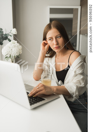Close up of young pretty woman drinking tea while break after typing and browsing online on laptop working on computer while sitting in cozy room alone. Distance learning and work with break for 98696892