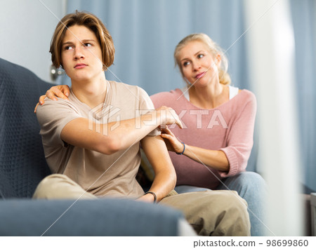 Upset and offended teenager sitting on sofa while mother soothing him 98699860
