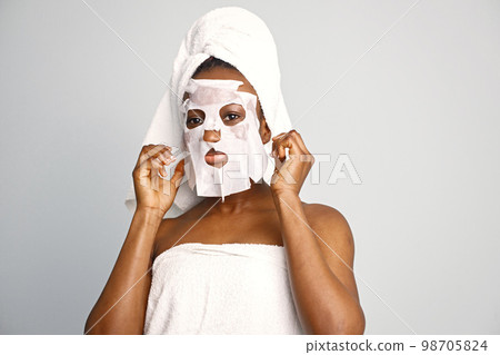 Black young woman standing isolated on white background with sheet mask on her face. Girl has her hair wrapped in a towel. Black woman enjoy after body treatment. 98705824