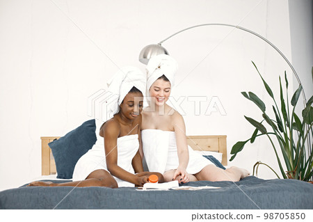 Two young women in white bathrobes sitting on a bed. Girls wearing white towels on their hair. Girls applying creams on the skin. Two young women in white bathrobes sitting on a bed. Girls wearing white towels on their hair. Girls applying creams on the skin. 98705850