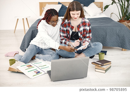 Two young women sitting on a floor near bed using a laptop at home. Black girl wearing eyeglasses and beige sweater, caucasian girl wearing plaid shirt. Girls preparing their homework. 98705895