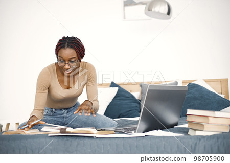 Young black woman sitting on bed using a laptop at home. Girl wearing eyeglasses and beige sweater. Girl preparing her homework. Young black woman sitting on bed using a laptop at home. Girl wearing eyeglasses and beige sweater. Girl preparing her homework. 98705900