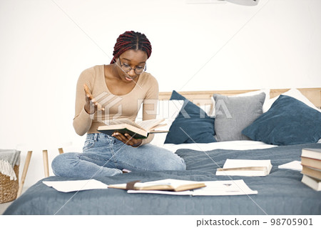 Young black woman sitting on bed reading a book at home. Girl wearing eyeglasses and beige sweater. Girl preparing her homework. 98705901