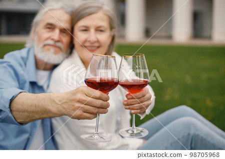 Focus on a two glasses of wine. Caucasian elderly couple sitting on a blanket on a grass in summer. Woman wearing white shirt and man blue one. 98705968