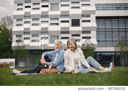 Beautiful caucasian elderly couple sitting on a grass in the park in summer. Man and woman sitting on blanket at the park drinking juice and sharing few precious memories. Woman wearing white shirt 98706018