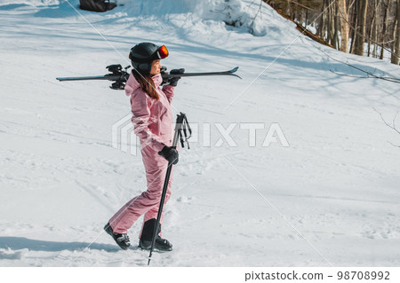 Winter Skiing. Ski portrait of woman alpine skier holdings skis wearing helmet, cool ski goggles, hardshell winter jacket and ski gloves on snow covered ski trail slope 98708992