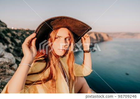 Portrait of happy young woman wearing summer black hat with large brim at beach on sunset. Closeup face of attractive girl with black straw hat. Happy young woman smiling and looking at camera at sea. 98709246