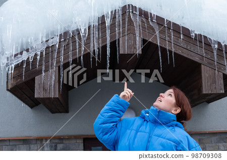 A frightened woman stands under a roof with large icicles. Danger in the winter season. 98709308