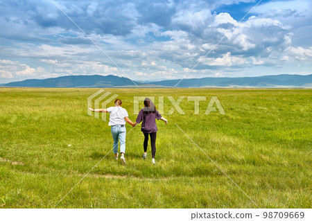Two girls run through the green grass of the Barguzin Valley in the Republic of Buryatia. View from the back. 98709669