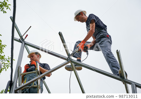 Workers installing metal beams for solar panels. Concept of renewable and ecological energy. Modern technology and innovation. Idea of environment safe. Men wearing workwear and working gloves Workers installing metal beams for solar panels. Concept of renewable and ecological energy. Modern technology and innovation. Idea of environment safe. Men wearing workwear and working gloves 98713011