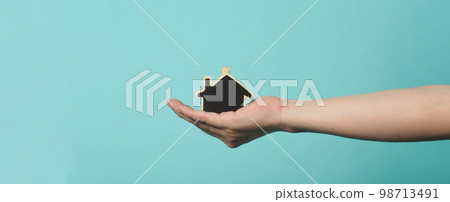 Small wood house in hands represent concepts such as home care family love real estate housing shelter insurance and mortgage. Hands holding small model house isolated on blue green studio background. Small wood house in hands represent concepts such as home care family love real estate housing shelter insurance and mortgage. Hands holding small model house isolated on blue green studio background. 98713491