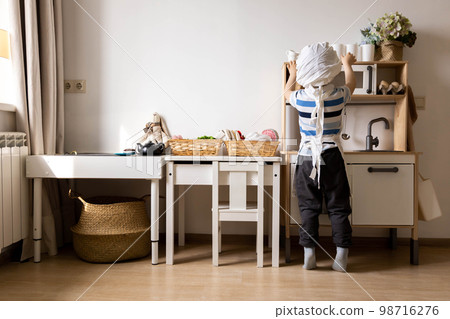 Cute little baby boy in chef hat and apron playing at childish kitchen cooking food back view Cute little baby boy in chef hat and apron playing at childish kitchen cooking food back view 98716276