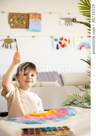 Confident cute male kid art drawing multicolored rainbow arch use paints and brush on table Confident cute male kid art drawing multicolored rainbow arch use paints and brush on table 98716319
