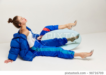 Portrait of beautiful woman in retro sports style trousers and down jacket posing with mirror over light background. Concept of weird people, fashion, creativity, aspiration 98716709