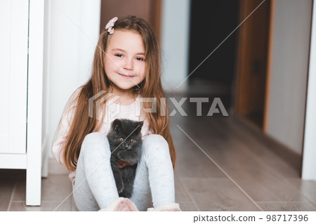 Smiling kid girl 4-5 year old holding small kitten sitting on floor in room. Looking at camera. Childhood. 98717396