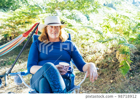 Adult woman relaxing, drinking tea while sitting in the travel chair near camper car vehicle and tents. Weekend trip activity. Female living on camper car and travel the world. Caravan car Vacation. Adult woman relaxing, drinking tea while sitting in the travel chair near camper car vehicle and tents. Weekend trip activity. Female living on camper car and travel the world. Caravan car Vacation. 98718912