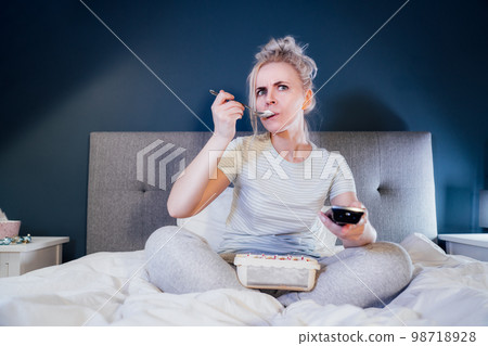 Shocked, stressed, surprised woman eating ice cream, holding a remote control while watching TV in bed. Scary horror movie or terrible news, scare in her eyes Overeating, Jamming of negative emotions. 98718928