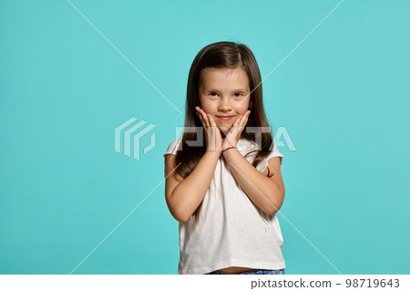 Close-up shot of a cute brunette little girl in a white blouse posing against a blue background. Close-up shot of a cute brunette little girl in a white blouse posing against a blue background. 98719643