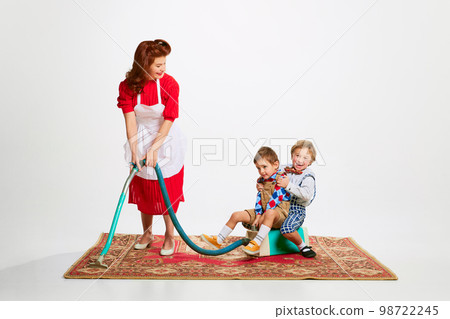 Domestic duties. Weekend cleaning. Beautiful woman cleaning carpet with vacuum cleaner with her playing kids, sons Domestic duties. Weekend cleaning. Beautiful woman cleaning carpet with vacuum cleaner with her playing kids, sons 98722245