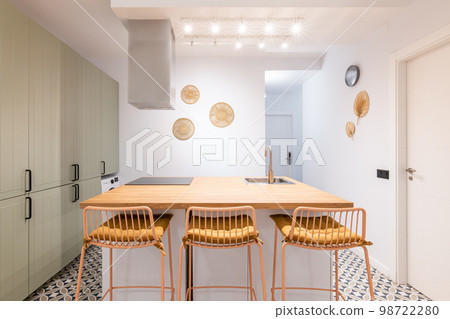Big kitchen island is surrounded by tall bar stools with soft yellow seat cushions. The table has a built-in ceramic hob and a sink with a faucet. 98722280