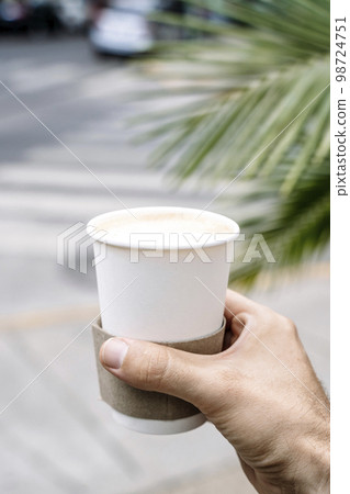 Takeaway coffee in a paper cup. A hand with a drink to go on the background of street. Paper cup mockup 98724751