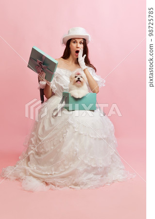 Present box with dog inside. Portrait of beautiful lady in white vintage dress posing over pink background. Concept of 19th century, fashion, comparison of eras Present box with dog inside. Portrait of beautiful lady in white vintage dress posing over pink background. Concept of 19th century, fashion, comparison of eras 98725785