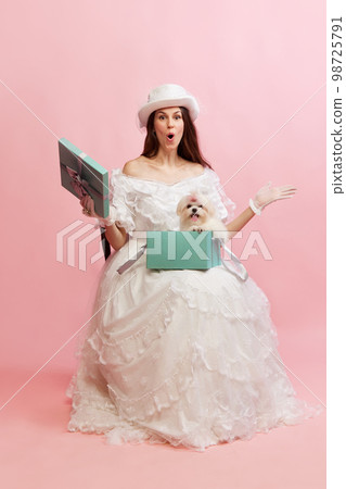 Surprise. Opening box with dog inside. Portrait of beautiful lady in white vintage dress posing over pink background. Concept of 19th century, fashion, comparison of eras Surprise. Opening box with dog inside. Portrait of beautiful lady in white vintage dress posing over pink background. Concept of 19th century, fashion, comparison of eras 98725791