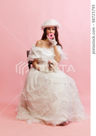 Eating donuts. Portrait of beautiful lady in white vintage dress posing with little dog over pink background. Concept of 19th century, fashion, comparison of eras Eating donuts. Portrait of beautiful lady in white vintage dress posing with little dog over pink background. Concept of 19th century, fashion, comparison of eras 98725795