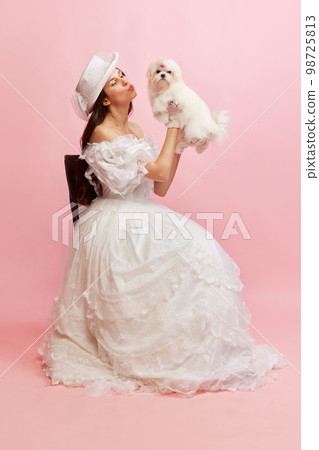 Portrait of beautiful lady, woman in white vintage dress posing with lovely cute dog over pink background. Concept of 19th century, fashion, comparison of eras Portrait of beautiful lady, woman in white vintage dress posing with lovely cute dog over pink background. Concept of 19th century, fashion, comparison of eras 98725813