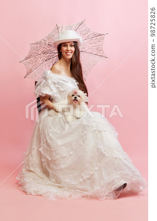 Woman and pet. Smiling. Portrait of beautiful lady in white vintage dress posing with dog over pink background. Concept of 19th century, fashion, comparison of eras Woman and pet. Smiling. Portrait of beautiful lady in white vintage dress posing with dog over pink background. Concept of 19th century, fashion, comparison of eras 98725826