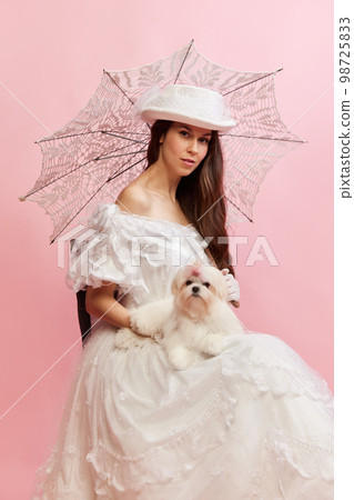 Woman and pet. Portrait of beautiful lady in white vintage dress posing with dog over pink background. Concept of 19th century, fashion, comparison of eras Woman and pet. Portrait of beautiful lady in white vintage dress posing with dog over pink background. Concept of 19th century, fashion, comparison of eras 98725833