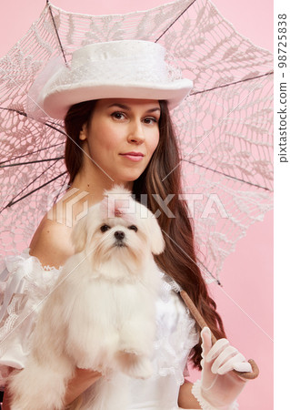 Family look. Portrait of beautiful lady, woman in white vintage dress with umbrella posing with cute dog over pink background. Concept of 19th century, fashion, comparison of eras Family look. Portrait of beautiful lady, woman in white vintage dress with umbrella posing with cute dog over pink background. Concept of 19th century, fashion, comparison of eras 98725838