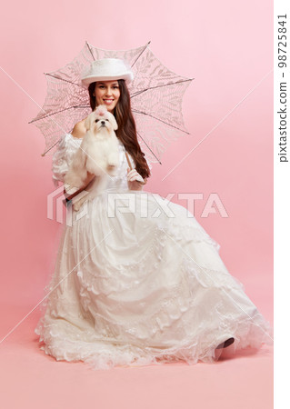 Portrait of beautiful lady, woman in white vintage dress posing with cute dog over pink background. Concept of 19th century, fashion, comparison of eras 98725841