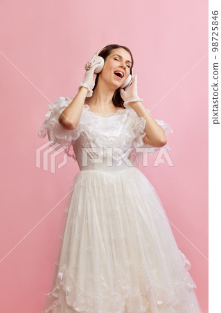 Portrait of beautiful lady in white vintage dress posing, listening to music in headphones over pink background. Concept of 19th century, fashion, comparison of eras Portrait of beautiful lady in white vintage dress posing, listening to music in headphones over pink background. Concept of 19th century, fashion, comparison of eras 98725846