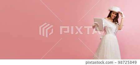 Modern devices. Video call. Portrait of beautiful lady in white vintage dress posing with tablet over pink background. Concept of 19th century, fashion, comparison of eras Modern devices. Video call. Portrait of beautiful lady in white vintage dress posing with tablet over pink background. Concept of 19th century, fashion, comparison of eras 98725849
