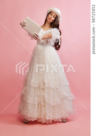 Sending kisses on online video call. Portrait of beautiful lady in white vintage dress posing with tablet over pink background. Concept of 19th century, fashion, comparison of eras 98725852