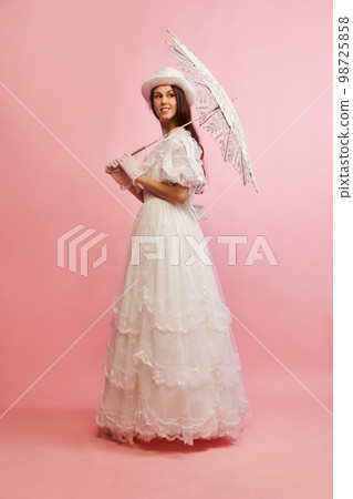 Tender femininity. Portrait of beautiful lady in white vintage dress posing with umbrella over pink background. Concept of 19th century, fashion, comparison of eras 98725858