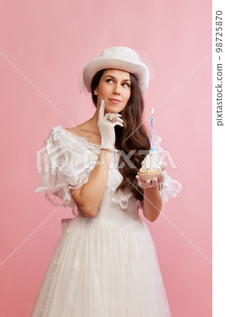 Making wishes. Portrait of beautiful lady in white vintage dress posing with birthday cupcake over pink background. Concept of 19th century, fashion, comparison of eras Making wishes. Portrait of beautiful lady in white vintage dress posing with birthday cupcake over pink background. Concept of 19th century, fashion, comparison of eras 98725870