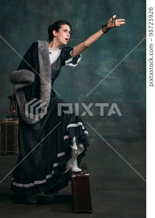 Getting off the train, arriving. Beautiful woman in image of Anna Karenina with retro suitcases over vintage dark green background. Concept of literature character, history Getting off the train, arriving. Beautiful woman in image of Anna Karenina with retro suitcases over vintage dark green background. Concept of literature character, history 98725926