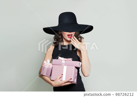 Happy surprised woman with pink gifts and in black wude broad brim hat on light grey background Happy surprised woman with pink gifts and in black wude broad brim hat on light grey background 98726212