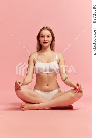 Young slim woman wearing white inner wear sitting in lotus yoga position over pink color background. Concept of natural beauty, body and skin care, healthy eating Young slim woman wearing white inner wear sitting in lotus yoga position over pink color background. Concept of natural beauty, body and skin care, healthy eating 98726290