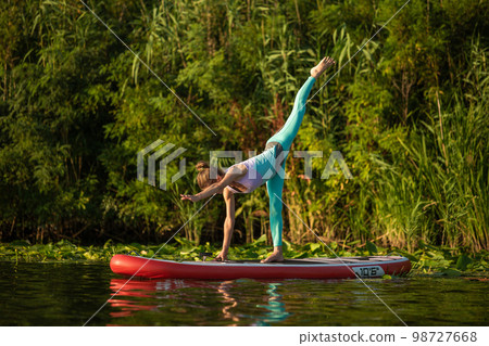 Photo of young woman doing hand stand on stand up paddle board. She wearing a leggings and top. 98727668