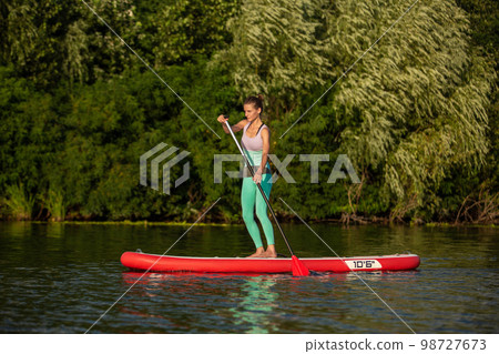 Young athletic woman doing fitness on a board with an oar on a lake. 98727673
